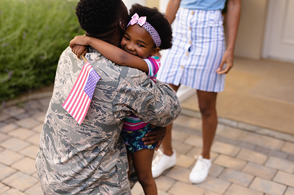 Army soldier embracing african american girl with usa flag at house entrance. family, bonding and patriotism, unaltered.